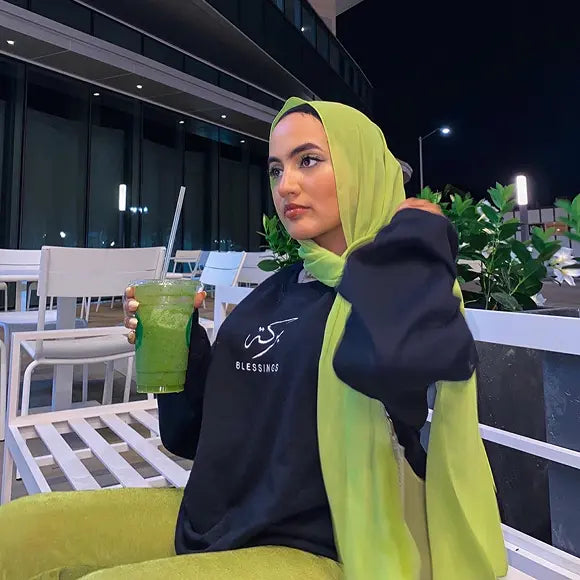 Muslim woman wearing SABR Blessings hoodie in black with green hijab holding a drink